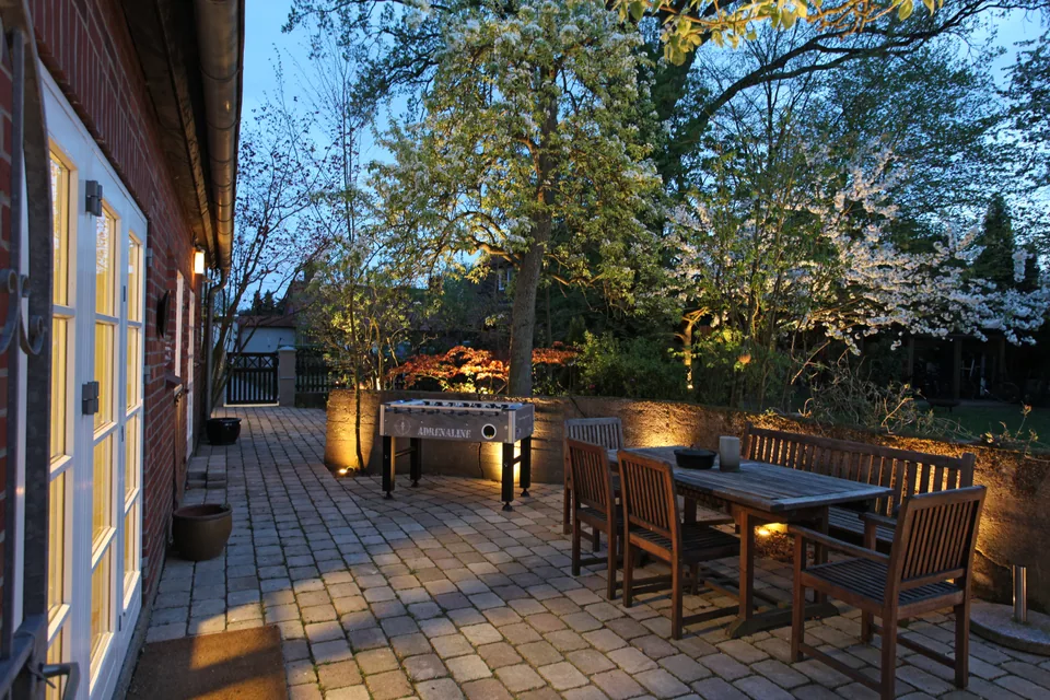 The terrace lit in the evening, with a wooden table, benches and table football beside the garden.