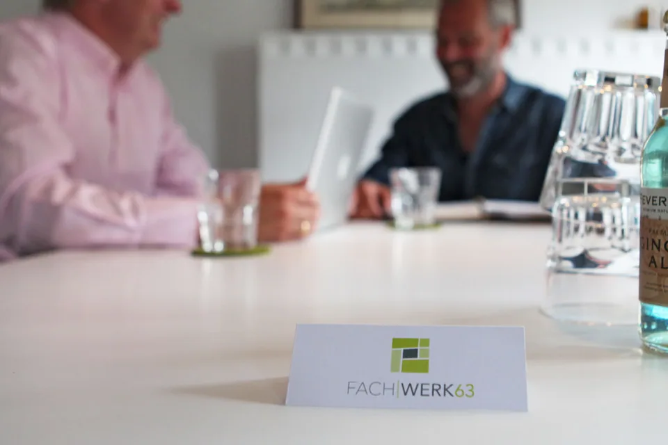 Two people at a meeting table, with a Fachwerk63 logo card in the foreground.