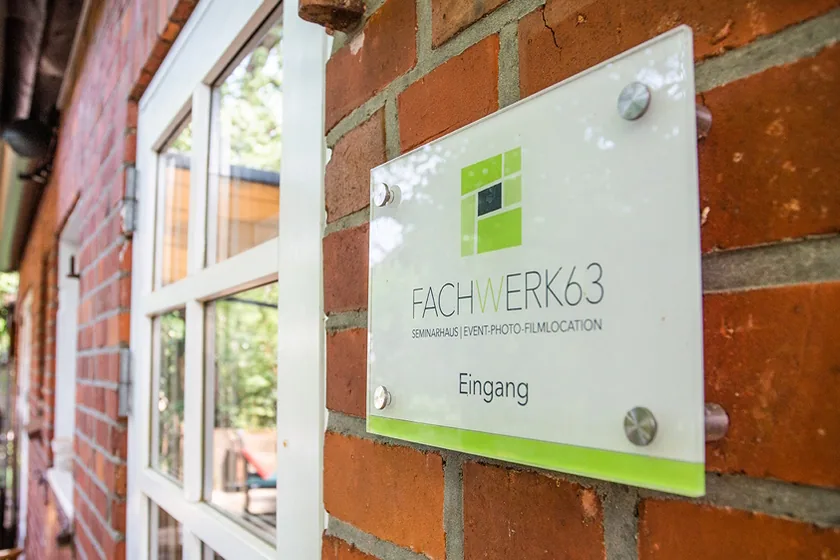 A Fachwerk63 entrance sign on a brick wall beside a glass door.